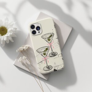 May include: A white iPhone case featuring a black and white illustration of two martini glasses with olives and pink ribbons. The case has a camera lens cutout and a white background.
