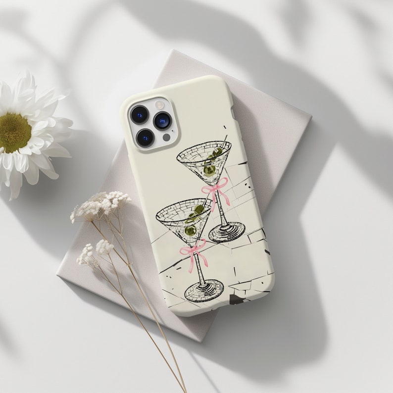 May include: A white iPhone case featuring a black and white illustration of two martini glasses with olives and pink ribbons. The case has a camera lens cutout and a white background.