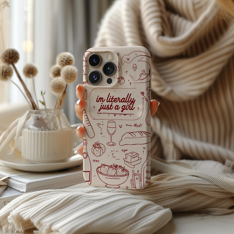 May include: A white phone case with a red and white design. The case features a variety of illustrations, including a glass of wine, a baguette, strawberries, and a heart. The text "im literally just a girl" is written in red on the case.