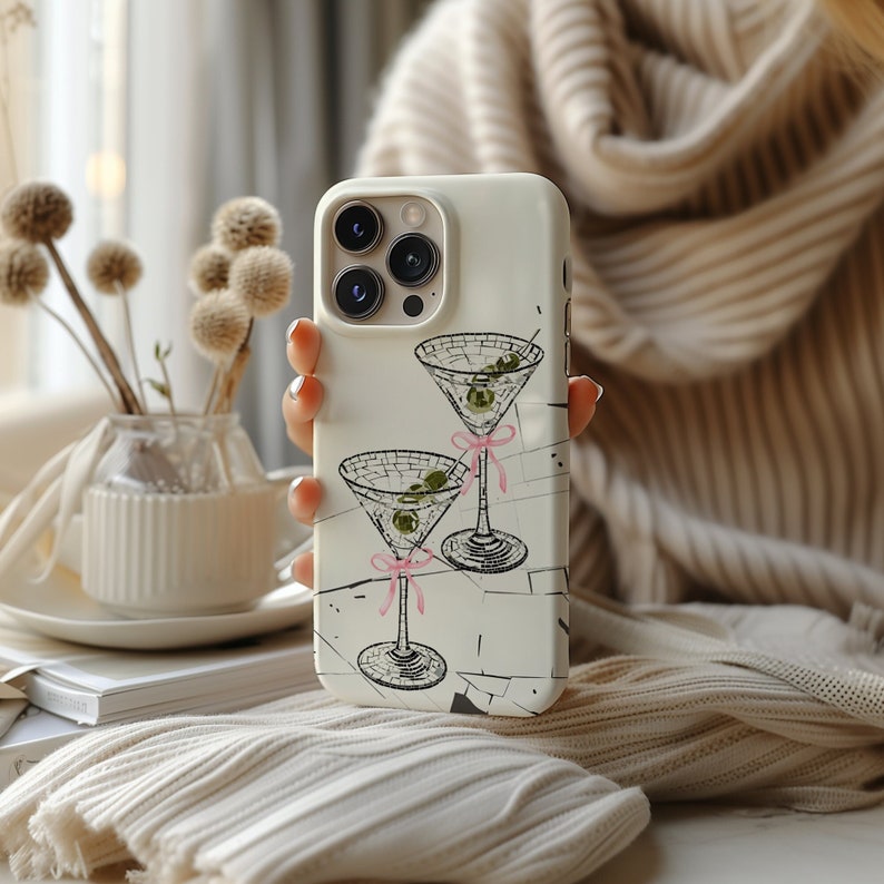 May include: White iPhone case featuring a design of two martini glasses with olives and pink ribbons. The case has a camera lens cutout and a white background.