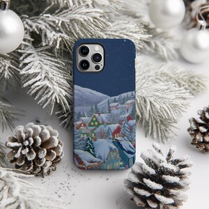 A smartphone case with a winter village scene printed on it, surrounded by pine cones and Christmas ornaments.