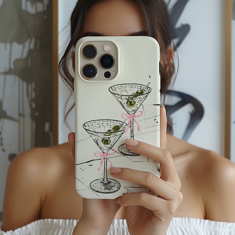 May include: White iPhone case featuring a design of two martini glasses with olives and pink ribbons. The case has a camera lens cutout and a white background.