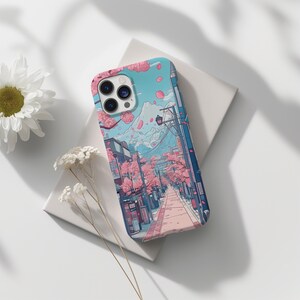 May include: A phone case with a Japanese street scene design, including cherry blossom trees, a mountain, and buildings. The case has a light blue background with pink and purple details. The phone case is on a white surface.