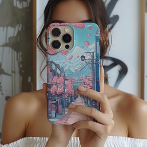 May include: A phone case with a Japanese-inspired illustration. The design depicts a street scene with cherry blossom trees, a mountain, and a blue sky. The case is a mix of pink, blue, and white.