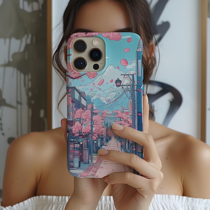 May include: A phone case with a Japanese-inspired illustration. The design depicts a street scene with cherry blossom trees, a mountain, and a blue sky. The case is a mix of pink, blue, and white.
