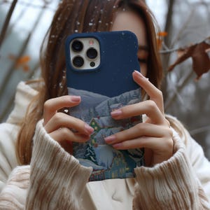 A person is holding a phone with a picture of a snowy village on the case, with snow falling around them.