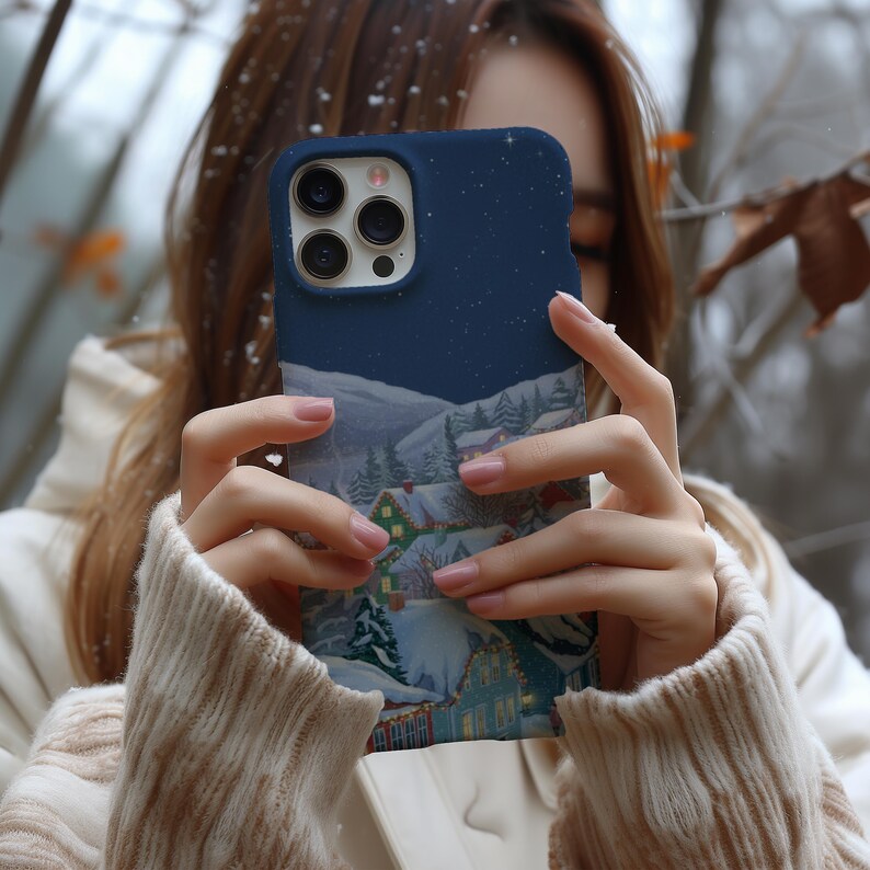 A person is holding a phone with a picture of a snowy village on the case, with snow falling around them.