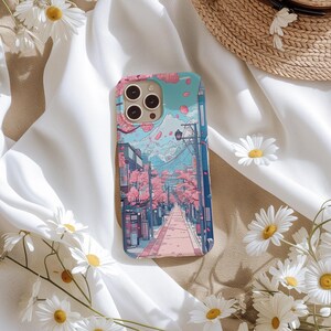 May include: A phone case depicting a Japanese street scene with cherry blossom trees, a mountain, and buildings. The case has a light blue background with pink and purple accents. The case has a camera cutout.