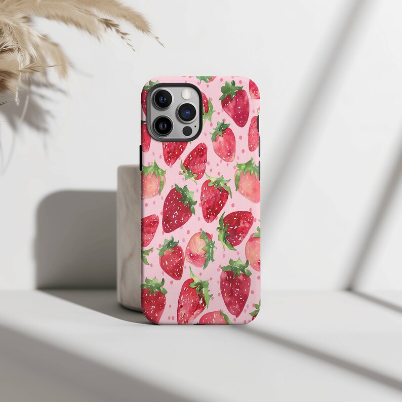 May include: A pink phone case featuring a pattern of red strawberries. The case has a cut-out for the camera lens.