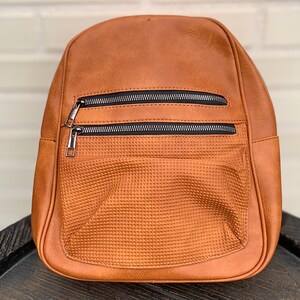 May include: A brown backpack with two zippered compartments and a textured front pocket. The backpack has a rounded top and adjustable straps. The zippers are black with silver pulls. The bag is made of a smooth, leather-like material.