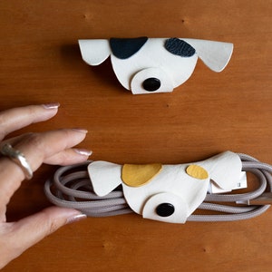 May include: Two white leather cord organizers shaped like dogs. One dog has black spots and the other has yellow spots. The dog with yellow spots is holding a gray cord.