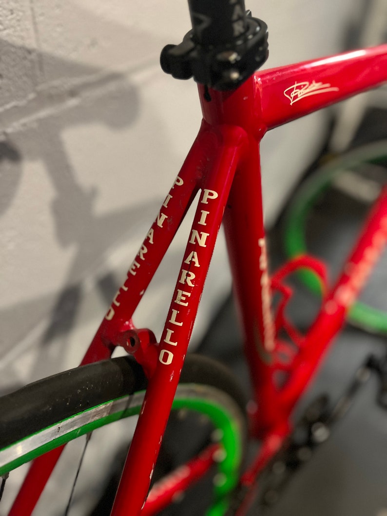 May include: Close-up of a red bicycle frame with white lettering that reads "PINARELLO". The bicycle has a green and silver rim.