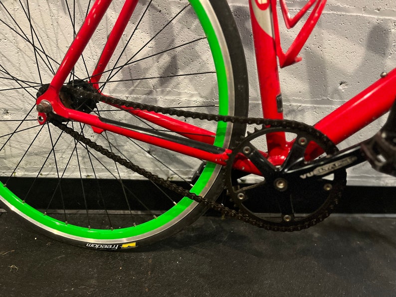May include: A red bicycle with a green rim and black chain. The bicycle has a black crankset and a black pedal.