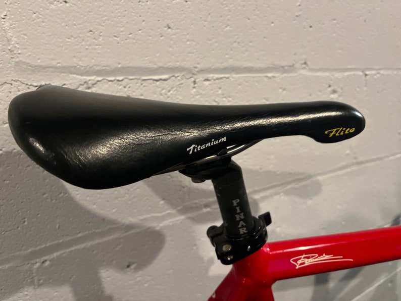 May include: A black bicycle seat with a white "Titanium" inscription and a gold "Flite" inscription. The seat is attached to a black seat post with a silver "Pinarello" inscription.