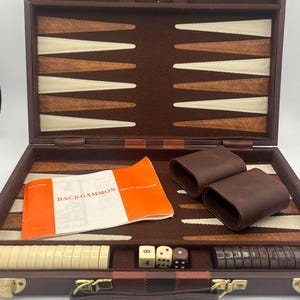 May include: Open backgammon set in a brown leather case with a handle and gold-colored clasps. The interior features a brown felt playing surface with tan and cream-colored triangles. Includes dice, dice cups, and a booklet labeled "Backgammon."