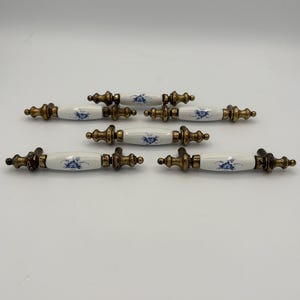 May include: A collection of seven antique cabinet pulls. Each pull features a white porcelain handle with a blue floral design and brass-toned metal hardware. The pulls are arranged on a white background.