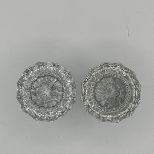 May include: Two clear, faceted glass knobs with a circular, floral-like design. The knobs are likely for a cabinet or drawer and have a vintage or antique aesthetic. The glass has a reflective quality.