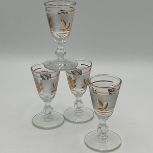 May include: Four clear glass cordial glasses with gold leaf and rim detailing. The glasses are stacked, with one glass on top of three others. The glasses have a frosted appearance and are set against a white background.