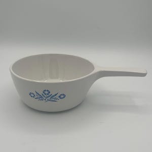 May include: White ceramic bowl with a handle, featuring a blue floral design on the side. The bowl has a rounded shape and a long, flat handle extending from the side. Ideal for serving soups or side dishes.