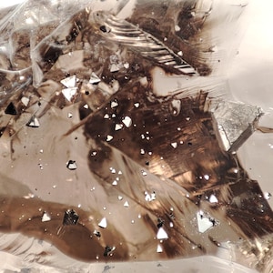 Könnte beinhalten: Abstraktes Bild einer durchscheinenden, kristallinen Struktur mit eingebetteten dunkelbraunen und silbernen Elementen. Verstreute, kleine, weiße und schwarze geometrische Formen sind in der Struktur aufgehängt und erzeugen einen dynamischen visuellen Effekt. Die Gesamtpalette ist neutral, mit Highlights.