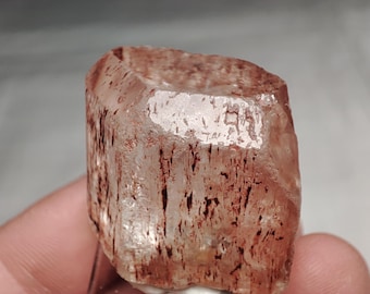 Op de afbeelding: Een helder, roodbruin kristal met een facetgeslepen bovenkant en een ruw, getextureerd oppervlak. Donkerdere rode insluitsels strepen verticaal door de doorschijnende steen. Het kristal wordt in een hand gehouden.