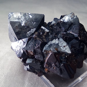 May include: A cluster of dark, metallic-looking crystals, possibly a mineral specimen. The crystals have sharp, geometric shapes and reflect light, creating a shimmering effect. The overall color is a deep, charcoal gray with hints of brown.