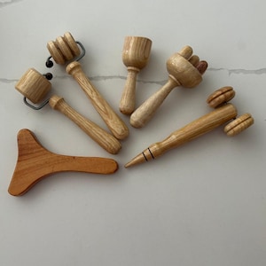 May include: A collection of wooden massage tools arranged on a white surface. The tools include rollers, mushroom-shaped massagers, and a pointed tool. The wood has a natural, light brown color. These tools are designed for massage and bodywork, promoting relaxation and muscle relief.