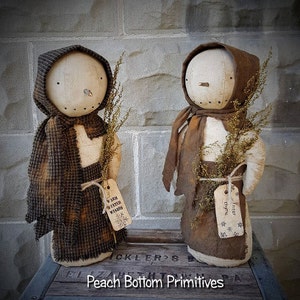May include: Two handmade primitive rag dolls dressed in brown fabric clothing and holding dried flowers. The dolls are standing on a wooden crate with the text "Peach Bottom Primitives". One doll has a tag that says "Warm Winter Wishes" and the other doll has a tag that says "Winter Thyme".
