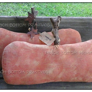 May include: Two rustic, peach-colored fabric pumpkins with wooden stems and metal star accents. One pumpkin has a tag that reads "Pumpkins 25¢". The pumpkins are displayed on a wooden surface, evoking a fall harvest theme.