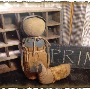 May include: A primitive rag doll with a brown and blue dress and a brown burlap skirt stands next to a small brown dog with a burlap collar. The doll and dog are in front of a wooden crate and a black sign that says "PRIM".