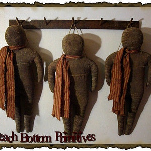 Op de afbeelding: Drie bruine stoffen poppen met rood-wit gestreepte sjaals die aan een houten rek hangen. De poppen zijn handgemaakt en hebben een rustieke, primitieve uitstraling. De tekst "Peach Bottom Primitives" is zichtbaar onderaan de afbeelding.
