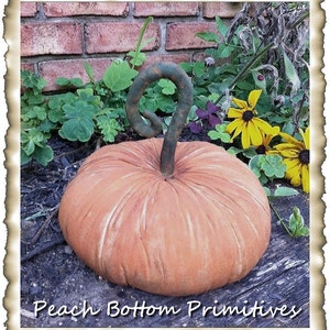 May include: A large, orange, fabric pumpkin with a green, twisted stem. The pumpkin is sitting on a wooden surface with a brick wall and green plants in the background. The text "Peach Bottom Primitives" is visible at the bottom of the image.