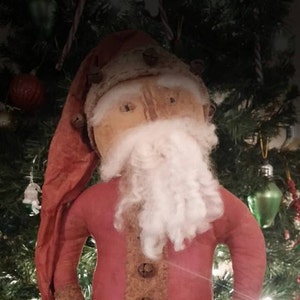 May include: A handmade rag doll Santa Claus with a red felt suit, white cotton beard, and a brown felt hat. The doll is standing in front of a Christmas tree.