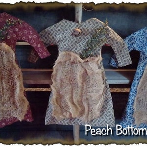 May include: Three handmade primitive style doll dresses with burlap skirts. The dresses are in red, blue, and green floral patterns. The dresses are hanging on a rustic wooden shelf. Peach Bottom Primitives.