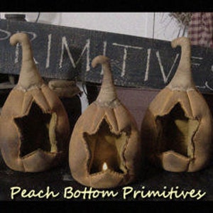 Op de afbeelding: Drie bruine stoffen pompoenen met een stervormige uitsparing en een kaars in de middelste pompoen. De pompoenen staan op een donker oppervlak met een houten bord waarop met witte krijt "PRIMITIVE" staat.