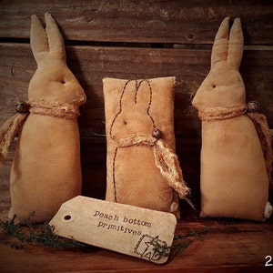 May include: Three handmade brown fabric bunny rabbits with a rustic, vintage look. The bunnies are sitting on a wooden surface with a tag that reads "Peach Bottom Primitives 2016".