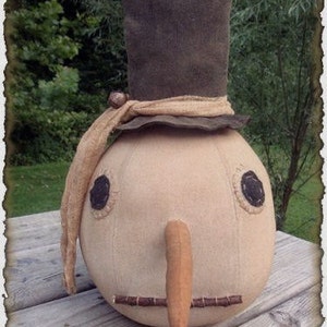 Peut inclure: Un poupon de neige primitif fait main avec un chapeau haut de forme en feutre marron, un nœud en toile de jute et un visage cousu avec un long nez en feutre orange. Le poupon est assis sur une surface en bois.