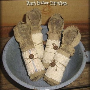 Op de afbeelding: Vier primitieve voddenpoppen, elk gewikkeld in stof en vastgebonden met touw, worden getoond in een blauwe metalen kom. Elke pop heeft een kleine bel eraan bevestigd. De poppen zijn gemaakt van jute en hebben een rustieke, handgemaakte uitstraling.