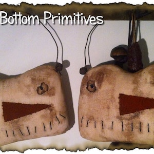 May include: Two handmade primitive fabric ornaments with stitched faces and red felt mouths. The ornaments are hanging from wire loops and have brown wooden beads at the top.