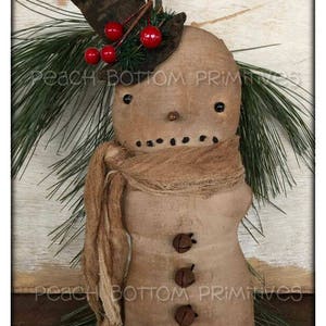 May include: A handmade snowman figure with a brown top hat, a beige scarf, and three brown button accents. The snowman's head is decorated with red berries and green sprigs. The figure is surrounded by pine needles.