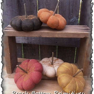 May include: Four handmade fabric pumpkins in various colors, including black, orange, red, and yellow, with a rustic wood and wire background. The pumpkins are all slightly different sizes and have a slightly distressed look. The text "Peach Bottom Primitives" is visible at the bottom of the image.