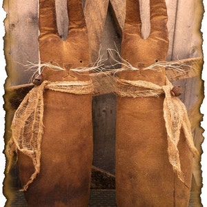 Può includere: Due bambole di coniglio primitive marroni con sciarpe di iuta e dettagli di baffi si trovano davanti a uno sfondo di legno rustico. Le bambole sono fatte a mano e hanno uno stile vintage e country.