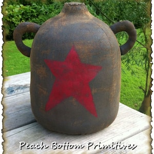 Könnte beinhalten: Ein rustikaler, dunkelbrauner Krug mit einem großen roten Stern auf der Vorderseite. Der Krug hat zwei Schlaufengriffe und eine strukturierte Oberfläche. Der Text "Peach Bottom Primitives" befindet sich am unteren Bildrand.