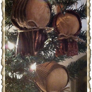 May include: Three handmade fabric ornaments in the shape of people with brown and white striped fabric and brown fabric. The ornaments are hanging on a green Christmas tree with white lights.