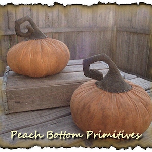 May include: Two orange fabric pumpkins with dark brown stems. The pumpkins are displayed on a wooden surface. The text "Peach Bottom Primitives" is at the bottom of the image.
