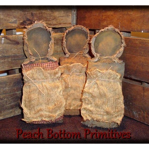 Op de afbeelding: Drie rustieke poppen met jute lijven, stoffen hoofden en touwaccenten. De poppen hebben een huiselijke uitstraling, met een mix van jute en stoffen met patronen. De poppen zijn gerangschikt voor een houten krat, met de tekst "Peach Bottom Primitives" onderaan.