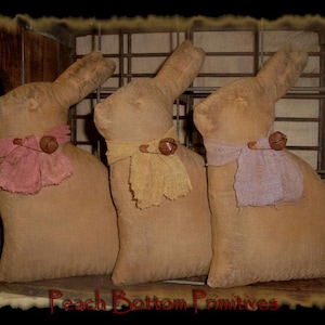 May include: Three handmade burlap bunny rabbits with fabric bows and bells. The bunnies are sitting on a wooden surface.