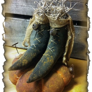 May include: Two rustic fabric boots with bells and burlap accents sit on top of a brown pumpkin. The boots are decorated with stitching and have a distressed look. The pumpkin is also distressed and has a rough texture. The image is titled "Peach Bottom Primitives 2014".
