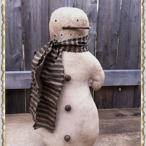 May include: A handmade fabric snowman with a striped scarf and three buttons. The snowman is standing on a wooden surface with a wooden fence in the background.
