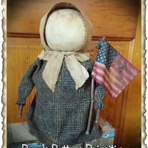 May include: A handmade rag doll with a brown and white plaid dress, a white bonnet, and a brown stick holding a small American flag. The doll is standing on a wooden box with the text "Peach Bottom Primitives" written on it.